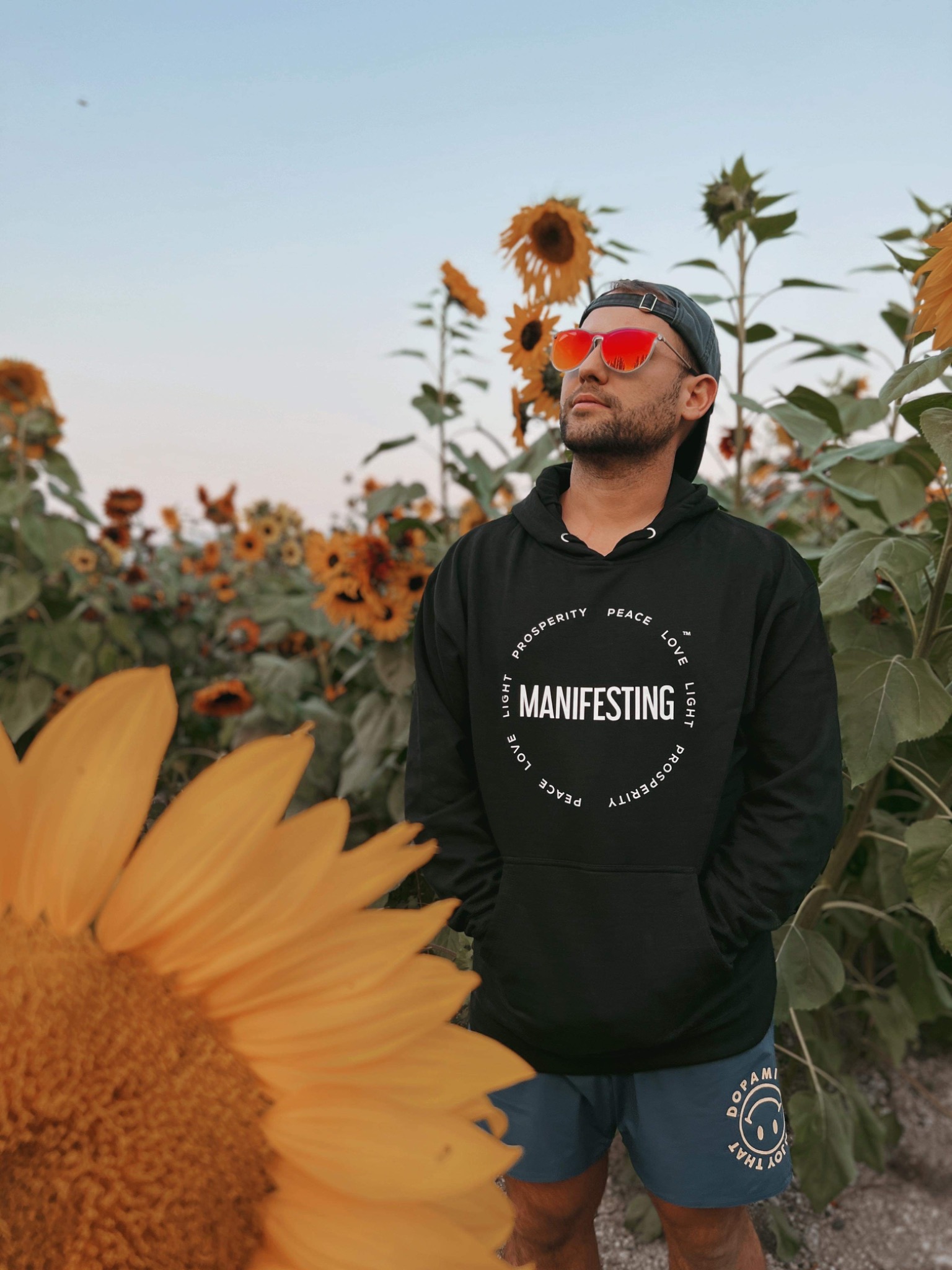 Man in a black Manifesting Circle hoodie standing in a sunflower field