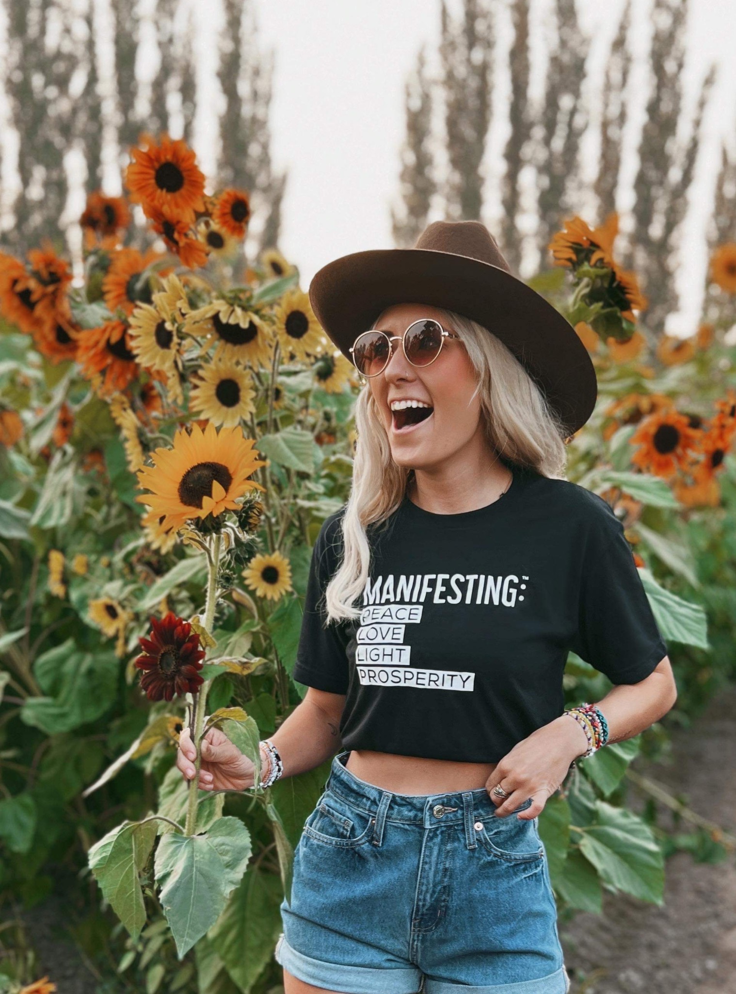 Woman laughing in a sunflower field wearing a black Manifesting tee